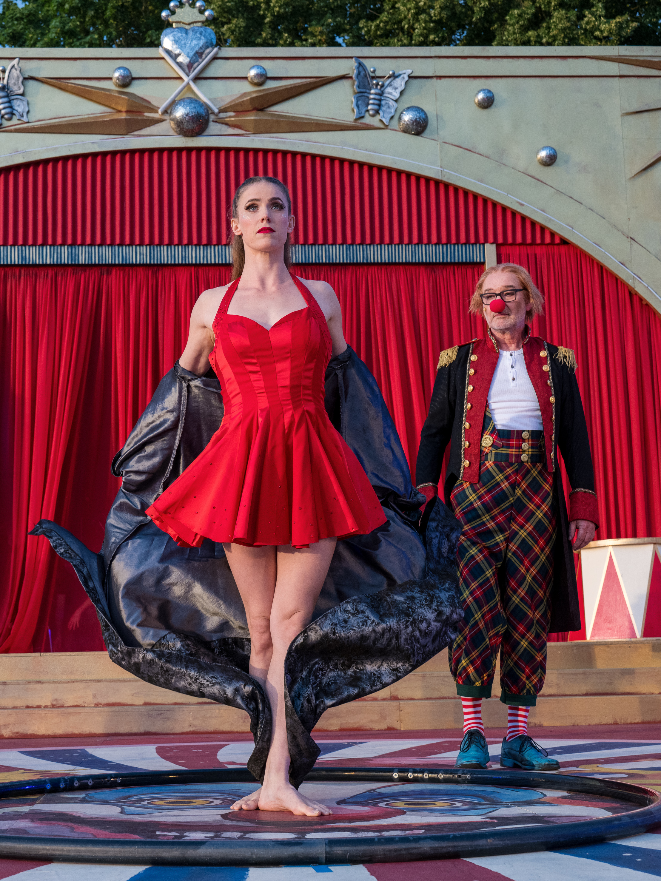 Eine Zirkusartistin im roten Kleid auf einer Bühne, im Hintergrund ein Clown