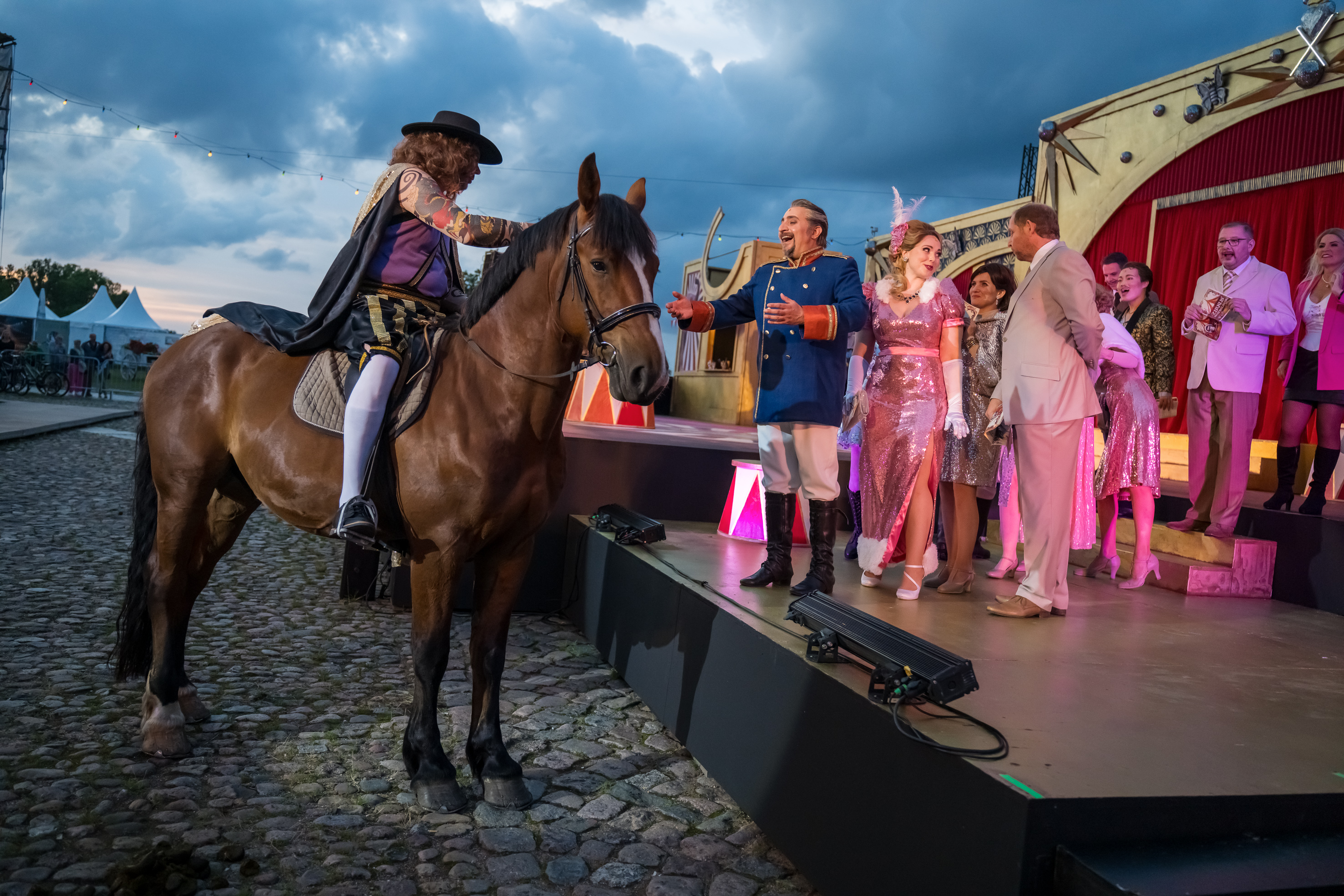Ein Pferd mit Reiter steht vor einer Bühne, darauf stehen mehrere Menschen hintereinander