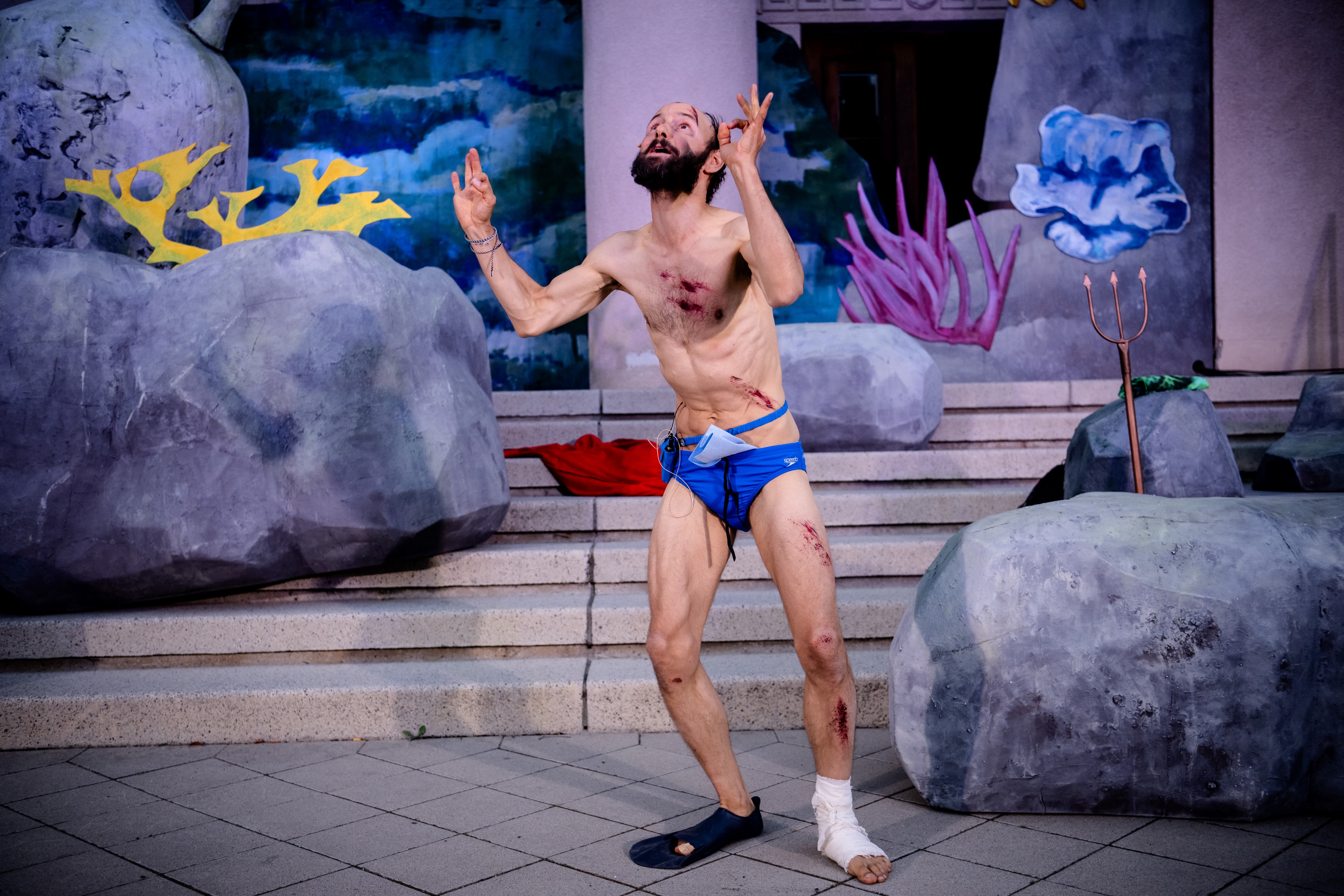 Ein Mann in Badehose und mit geschminkten Wunden am Körper blickt zum Himmel empor