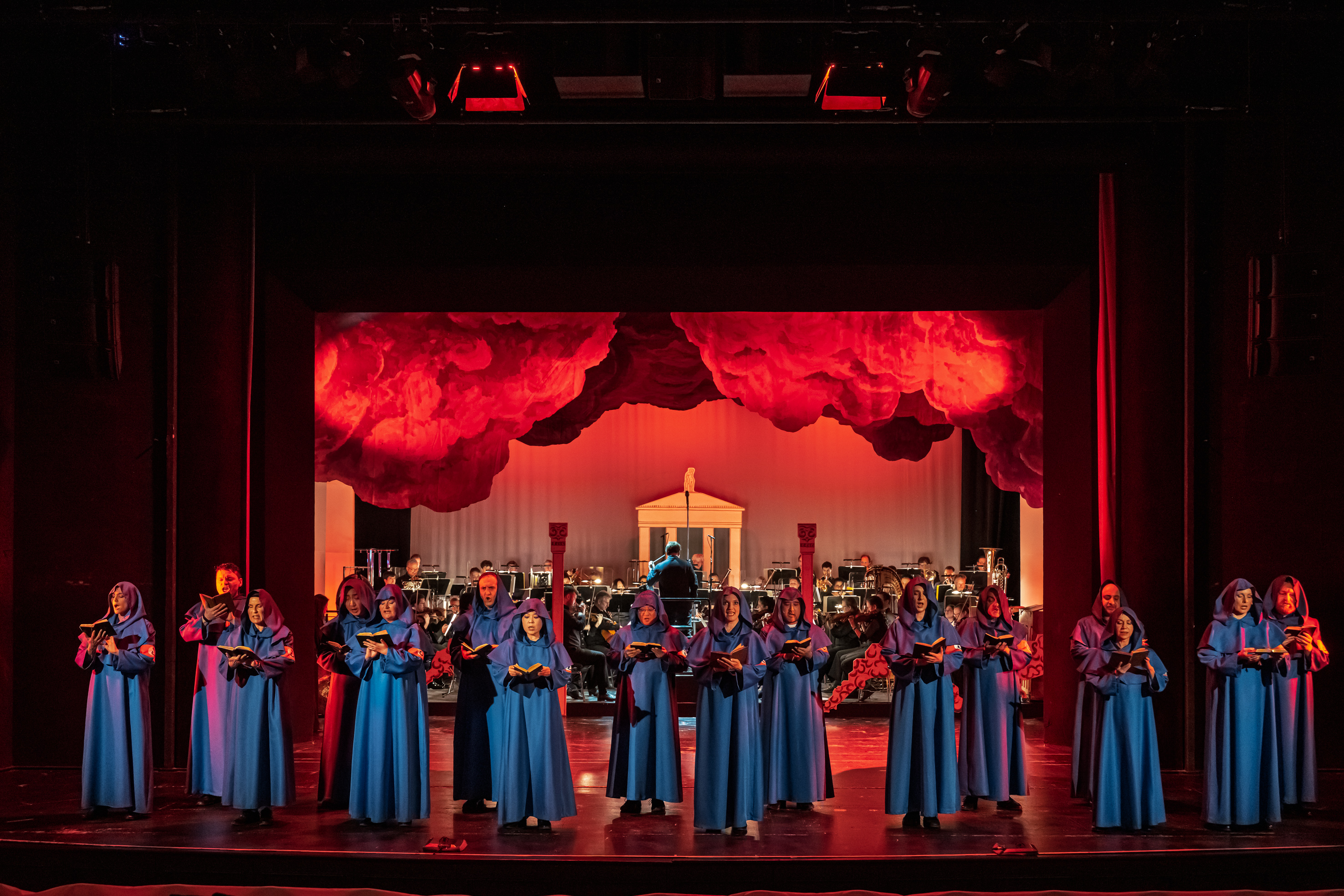 Opernchor in blauen Mönchskutten in rotem Lichtkegel im HIntergrund ein Orchester