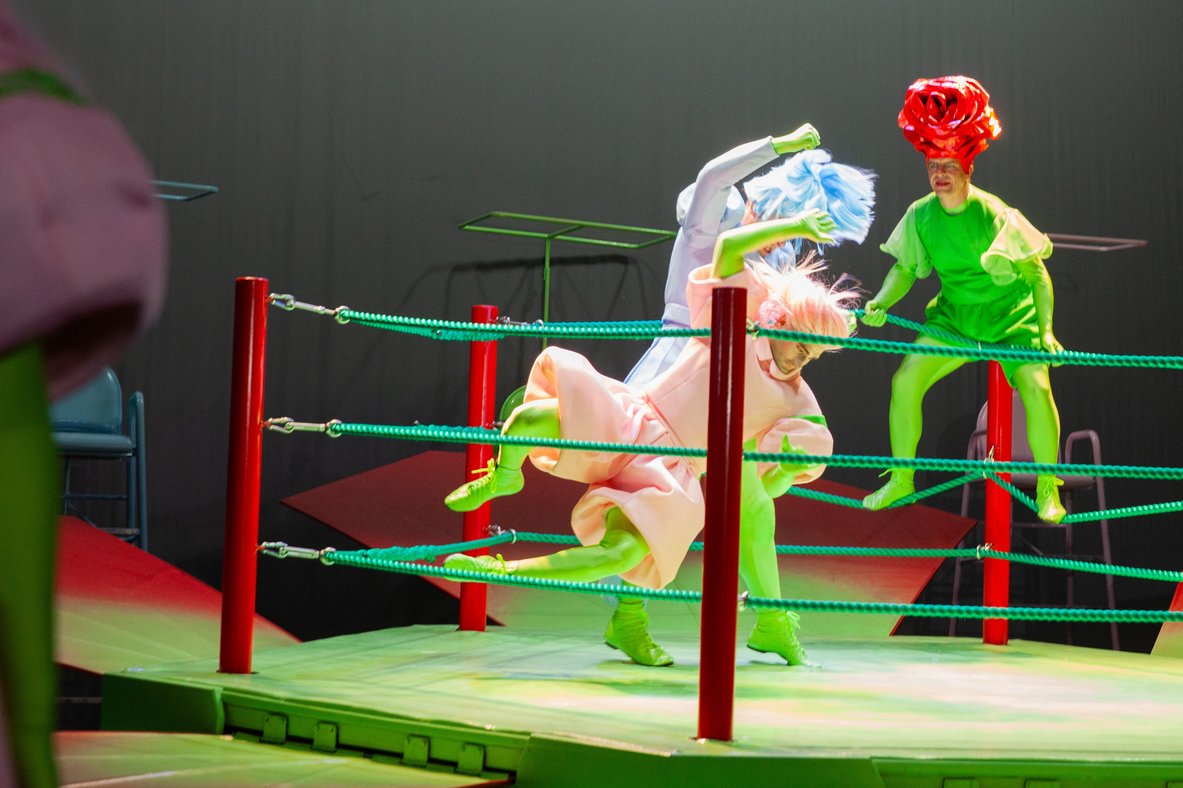 Zwei Personen in einem Ringkampf, eine dritte Person mit einer Rose auf dem Kopf schaut zu