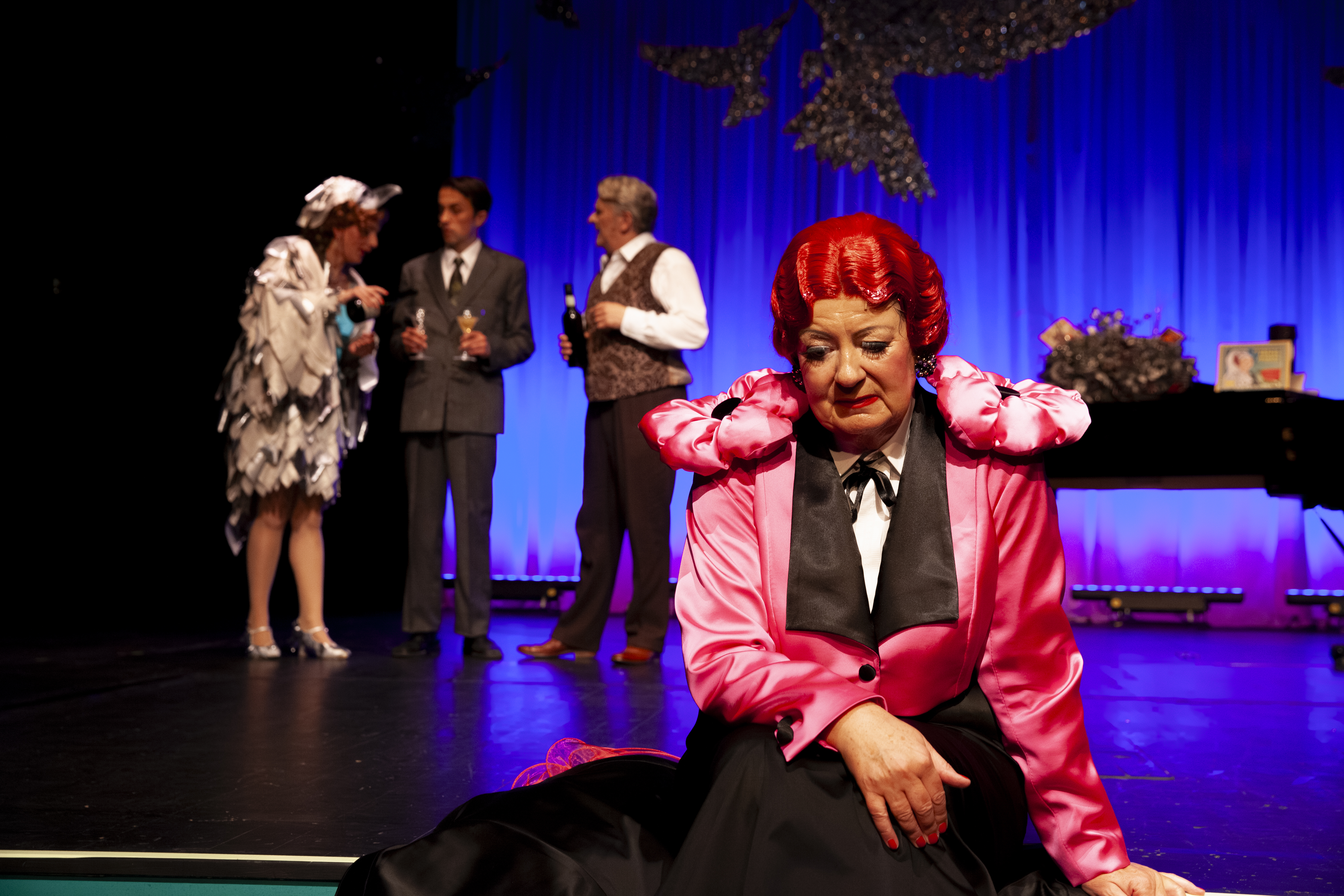 Eine Frau mit Riten Haaren sitzt am Boden und scheint traurig zu sein. Im Hintergrund sieht man drei Personen, die etwas trinken und sich unterhalten. Rechts von ihnen steht ein Piano.