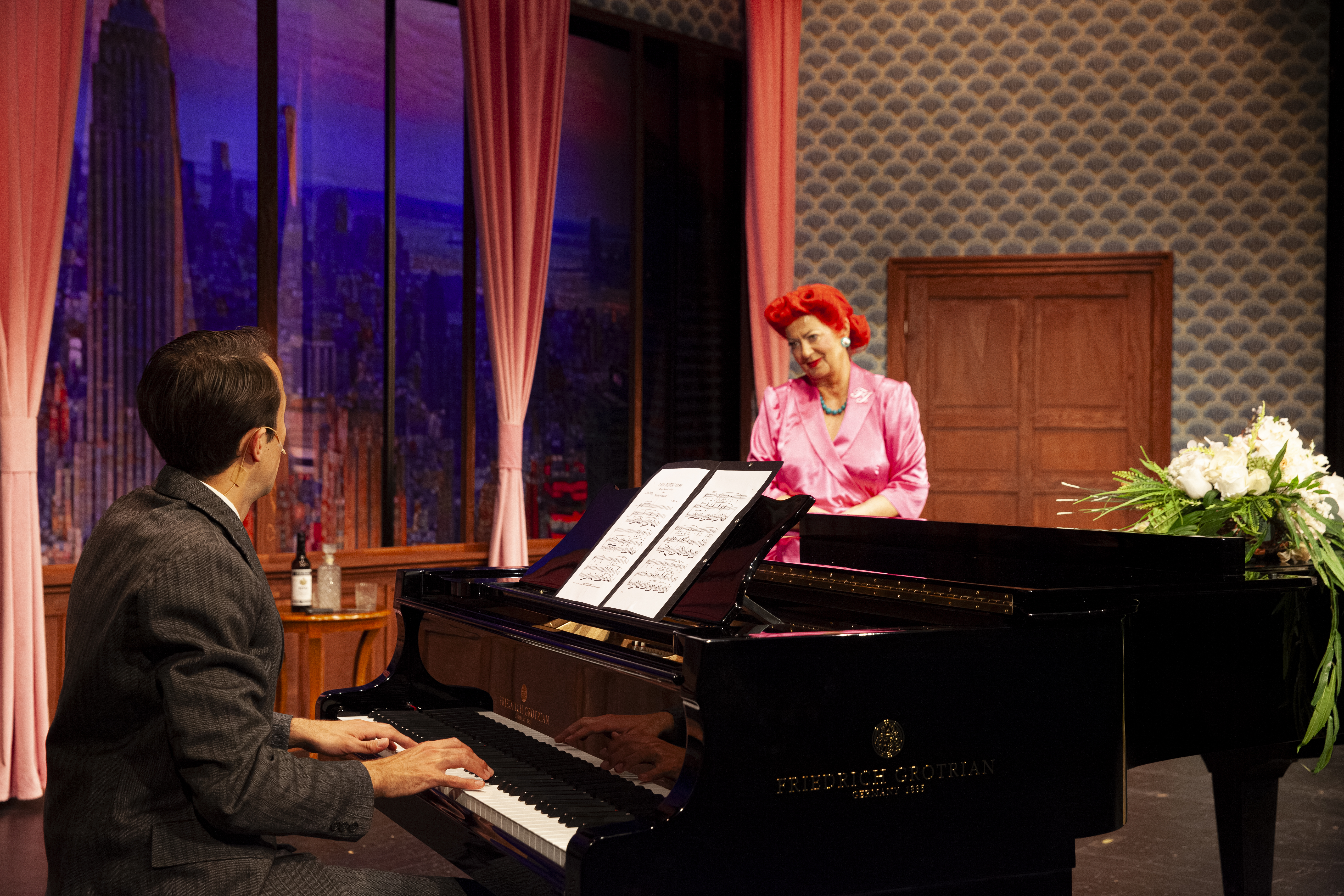 Ein Mann sitzt am Piano. Er trägt einen Anzug. Eine Frau mit roten Haaren lehnt am Piano. Sie wird gleich singen.