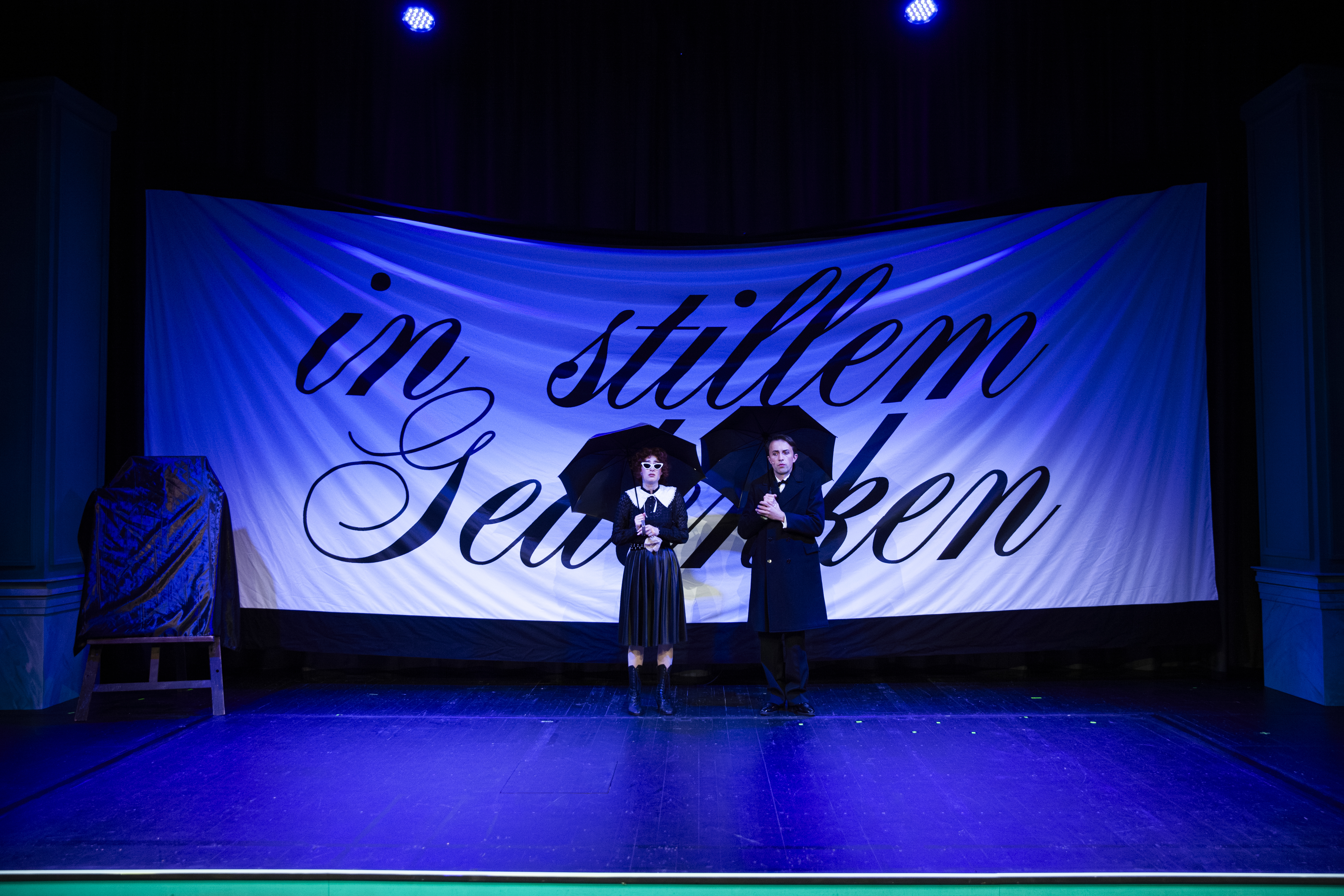 2 Personen in schwarz gekleidet mit Regenschirmen in der Hand stehen vor einem Banner. Auf dem steht: in stillem Gedenken.